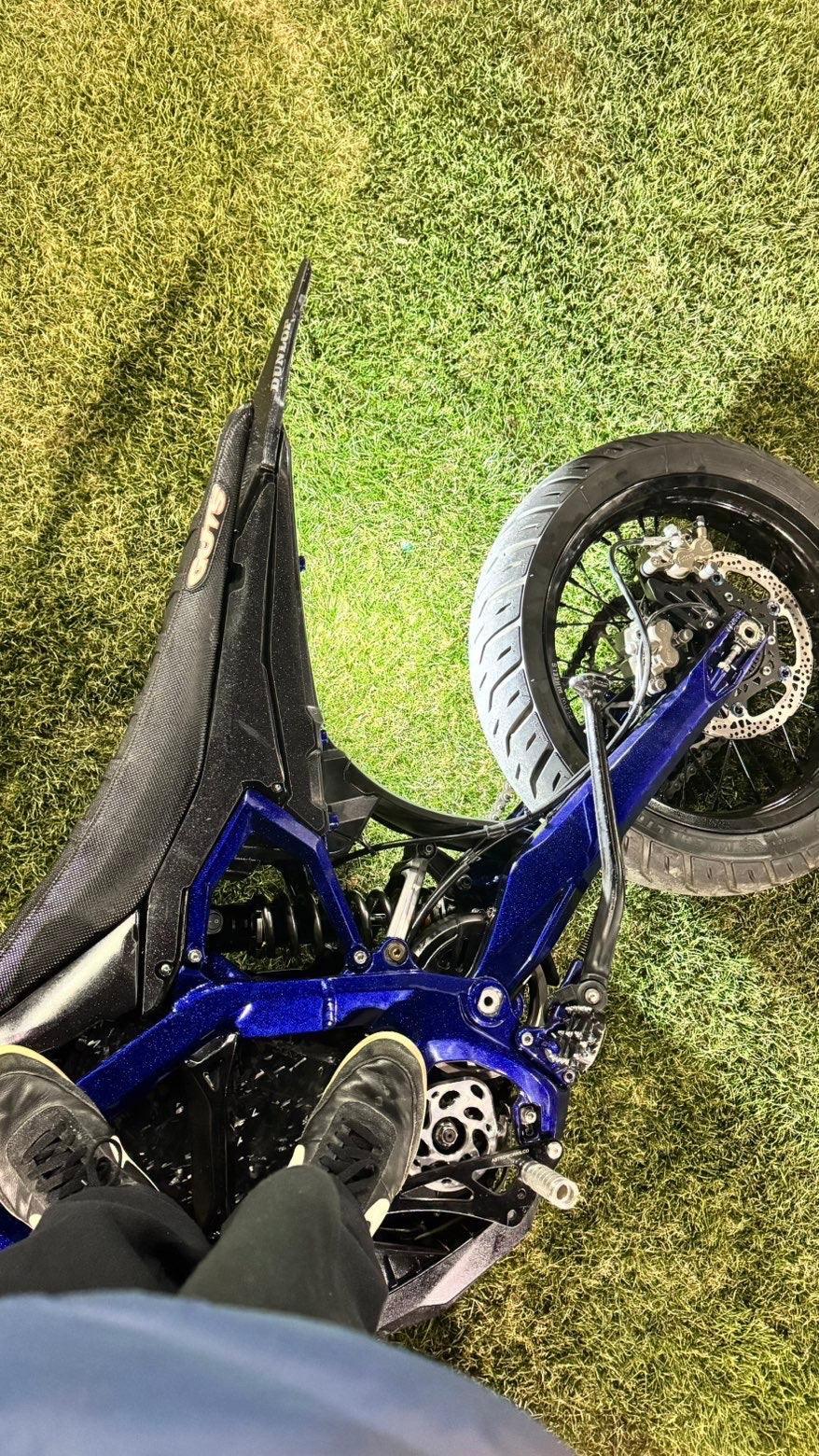 Rider's downward view of a blue bike with a polished motor cover beside the footpeg, parked on grass.