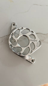 Close-up of a polished Riot motor cover on a light surface, showing its central opening, curved cutouts and engraved RIOT logo.