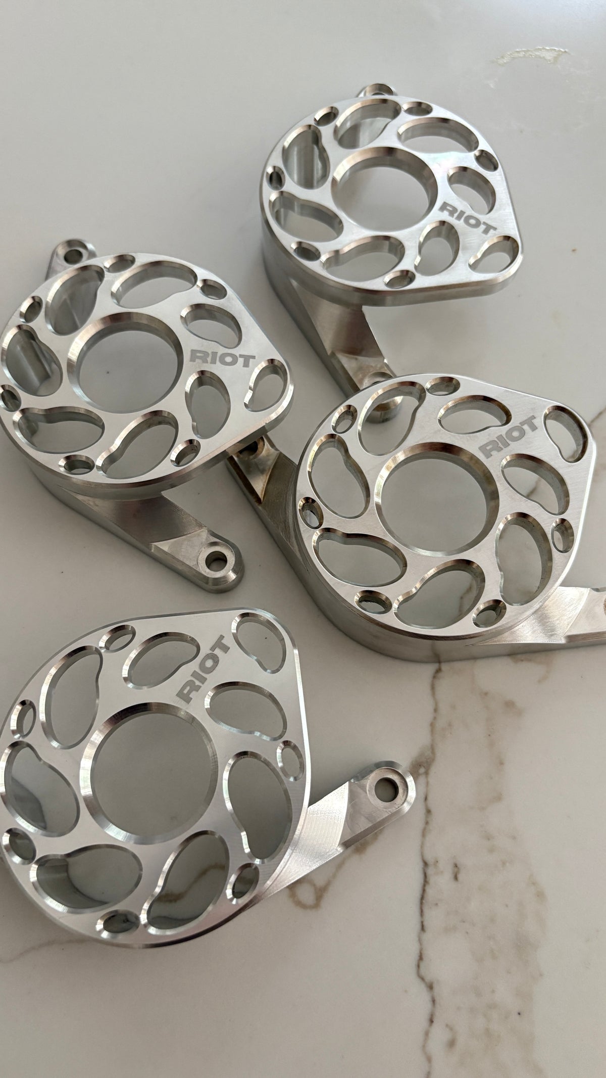 Four polished Riot motor covers arranged on a light stone-patterned surface, showing their circular cutouts and mounting tabs.