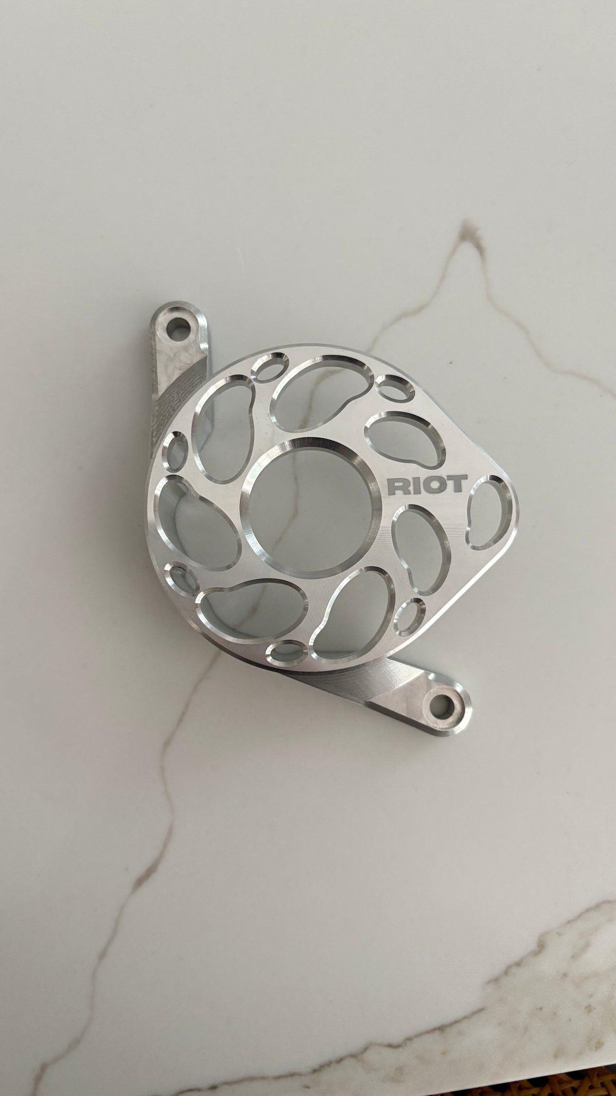 Overhead view of a polished Riot motor cover with a central opening and curved cutouts on a light stone-patterned surface.